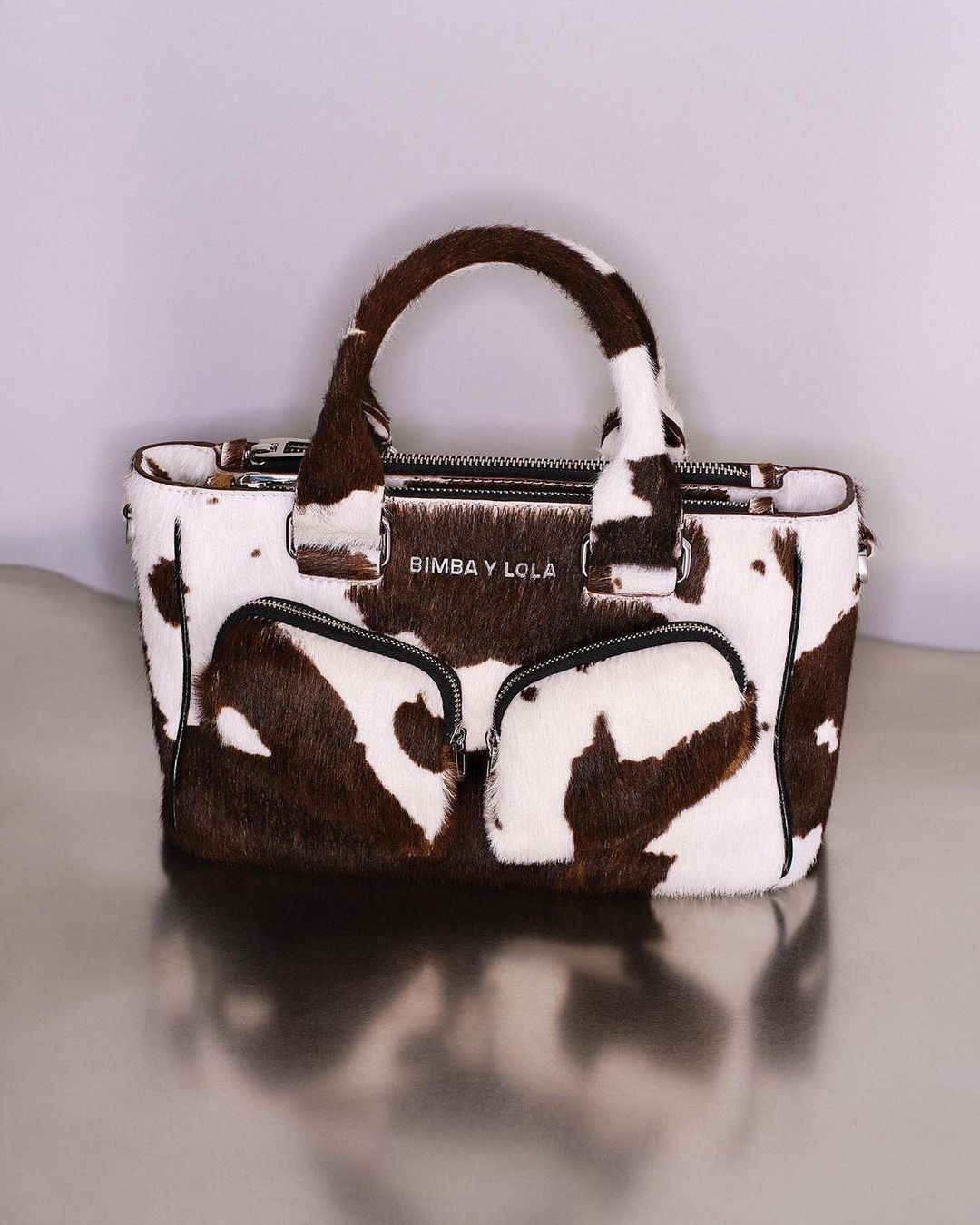 Fashionable handbag with cowhide print, featuring top handles and zippered compartments, set against a neutral backdrop in a studio setting.