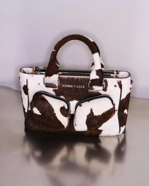 Fashionable handbag with cowhide print, featuring top handles and zippered compartments, set against a neutral backdrop in a studio setting.