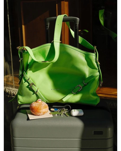 Lime green leather tote bag sitting on top of a gray hard shell suitcase next to a glazed donut, keys, and wireless earbuds.