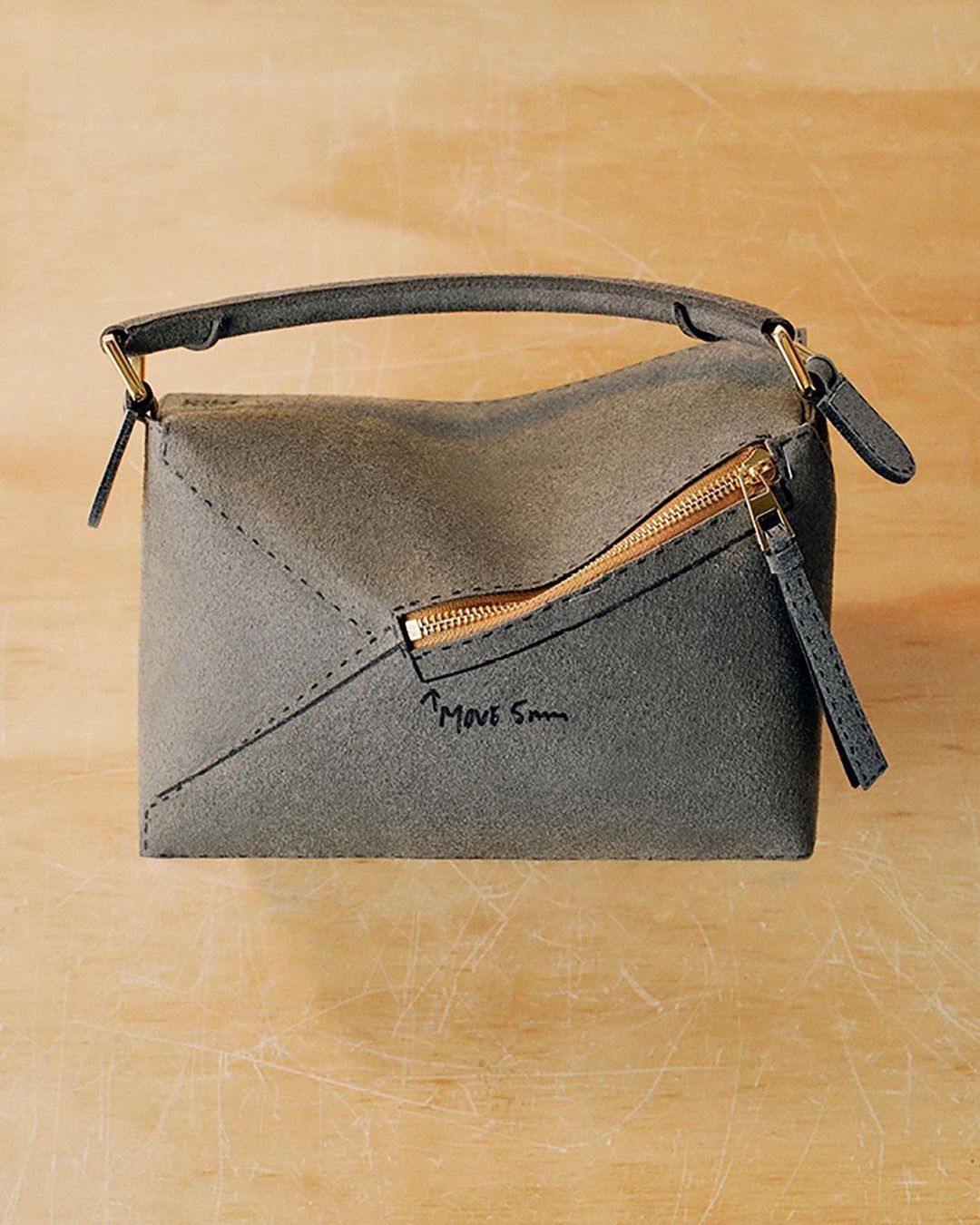 Gray suede envelope-style handbag with a top handle and a gold zipper detail, presented on a light brown surface.