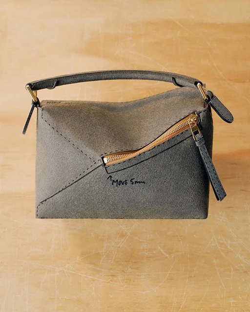 Gray suede envelope-style handbag with a top handle and a gold zipper detail, presented on a light brown surface.