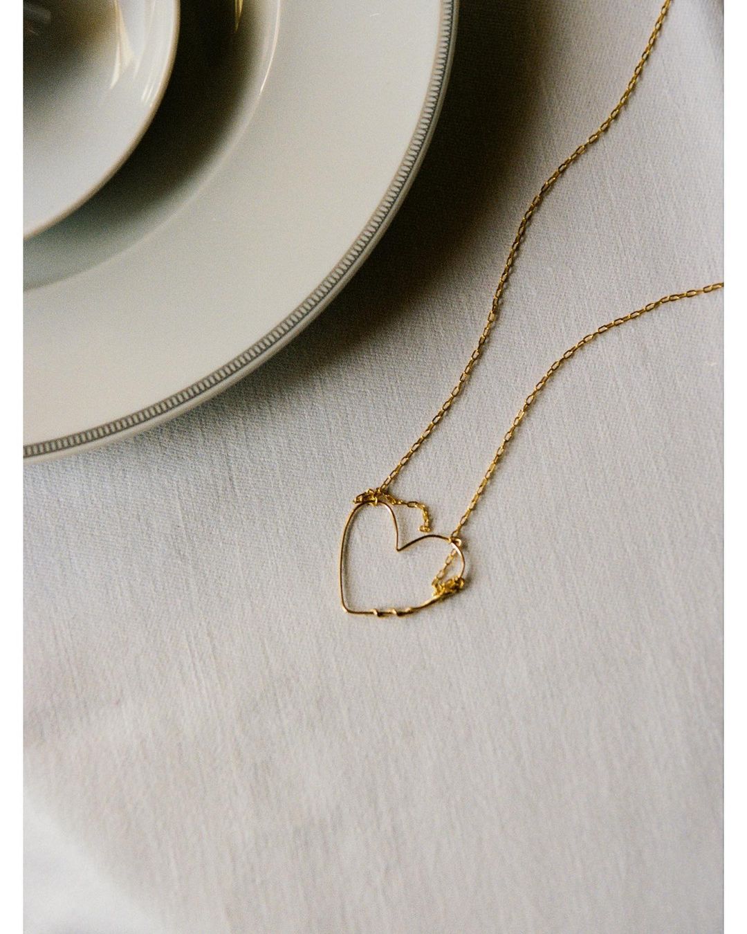 Gold necklace with a heart-shaped pendant on a white tablecloth next to white ceramic plates, showcasing jewelry and table setting.