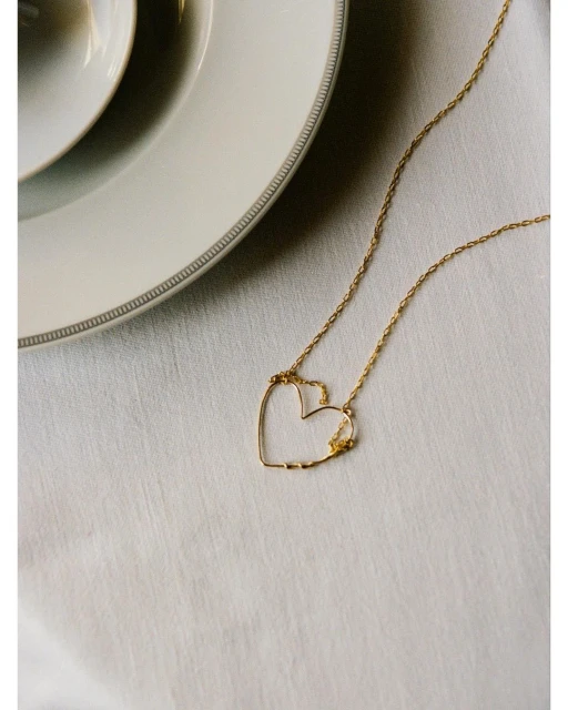 Gold necklace with a heart-shaped pendant on a white tablecloth next to white ceramic plates, showcasing jewelry and table setting.