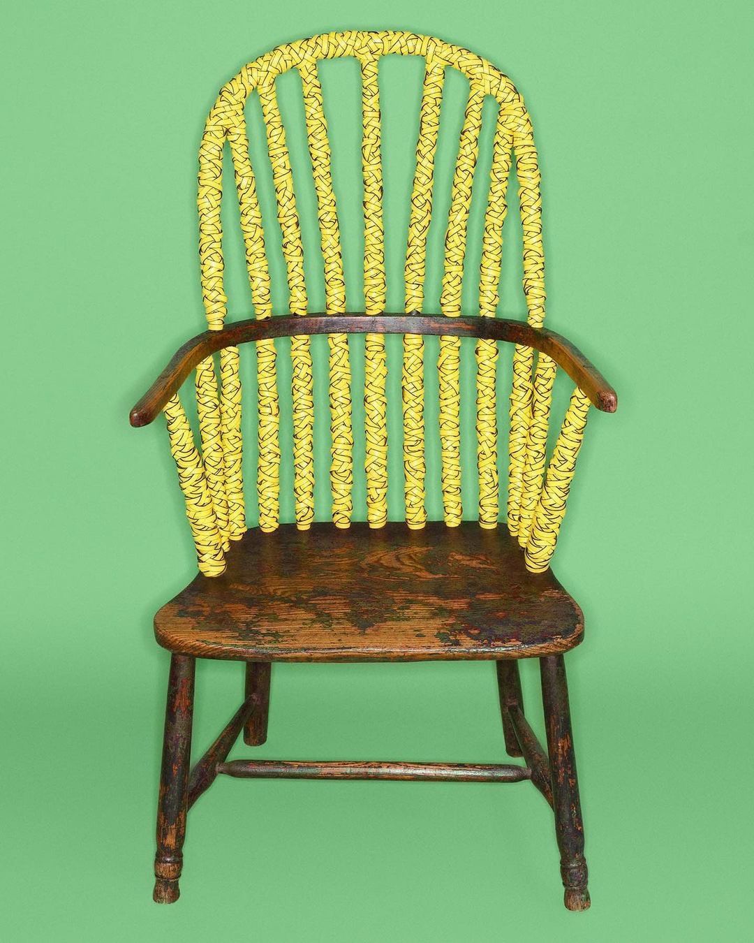 Antique Windsor chair featuring a painted yellow backrest with black rope detailing, wooden seat, and dark legs, set against green backdrop.