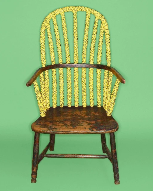 Antique Windsor chair featuring a painted yellow backrest with black rope detailing, wooden seat, and dark legs, set against green backdrop.