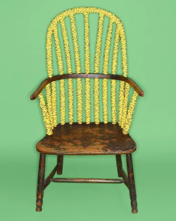 Antique Windsor chair featuring a painted yellow backrest with black rope detailing, wooden seat, and dark legs, set against green backdrop.