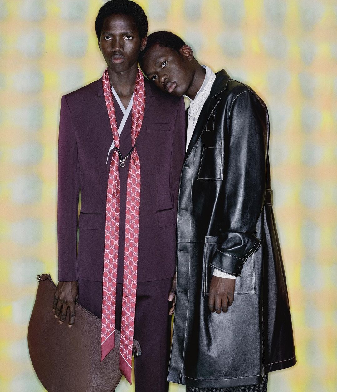 Two men stand in front of patterned yellow backdrop, one in a suit with a Gucci scarf, the other in a leather coat.