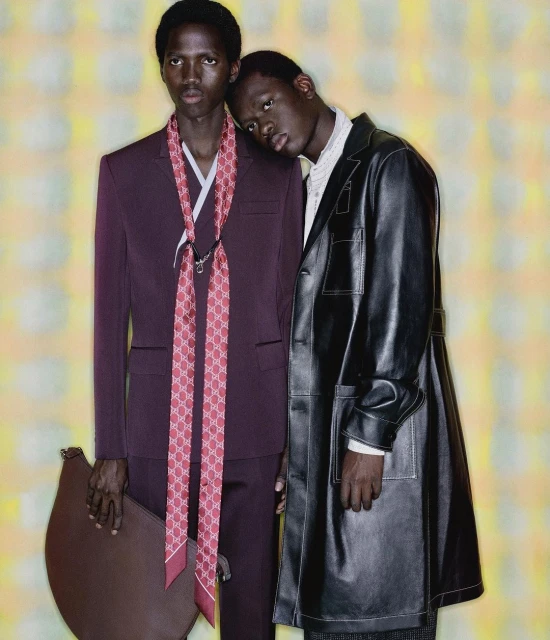 Two men stand in front of patterned yellow backdrop, one in a suit with a Gucci scarf, the other in a leather coat.