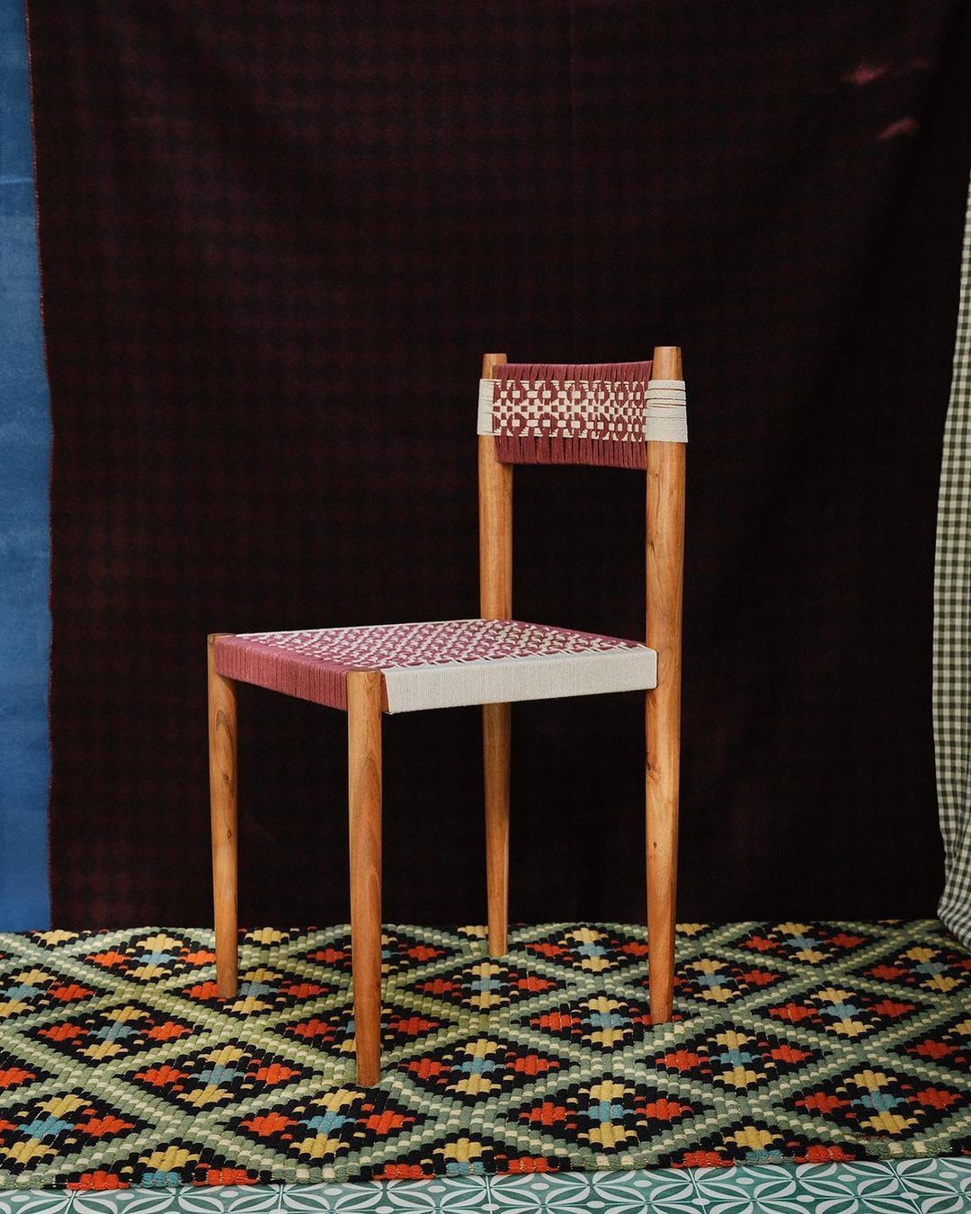Studio shot of a chair with a woven seat and backrest on a patterned rug in front of a backdrop with various colors and patterns.