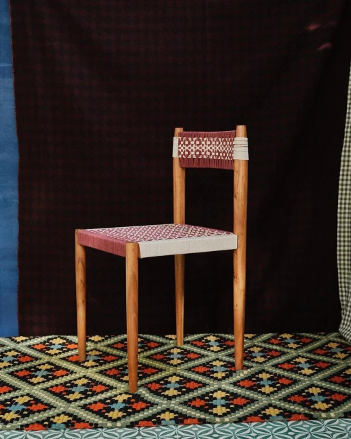 Studio shot of a chair with a woven seat and backrest on a patterned rug in front of a backdrop with various colors and patterns.