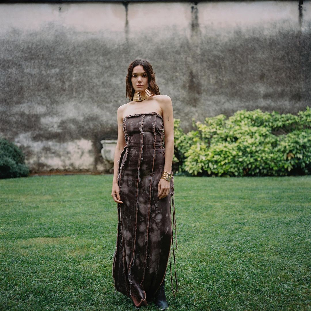 A woman models a strapless brown dress and gold jewelry on a green lawn against a weathered wall in a medium outdoor shot.