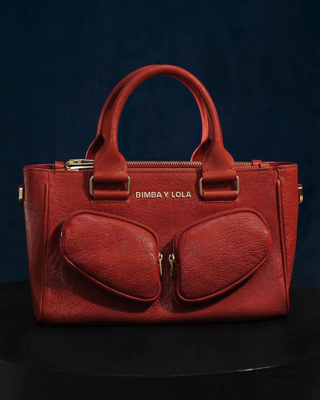 Red Bimba y Lola handbag featuring a textured leather-like material, gold hardware, and a structured design on a dark surface.