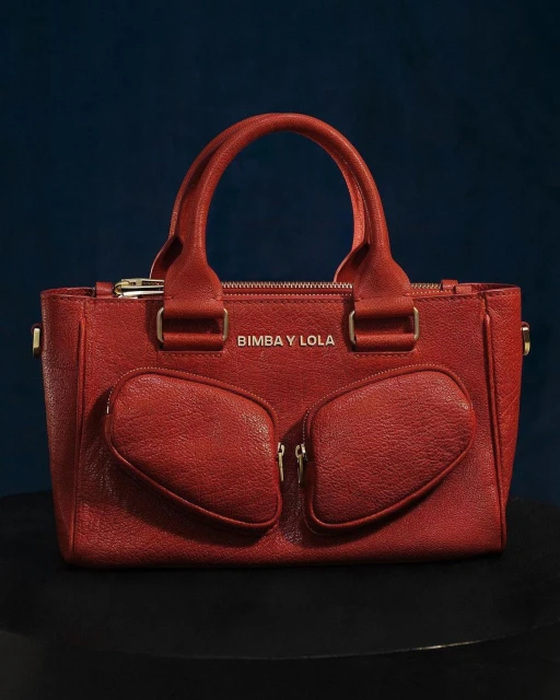 Red Bimba y Lola handbag featuring a textured leather-like material, gold hardware, and a structured design on a dark surface.