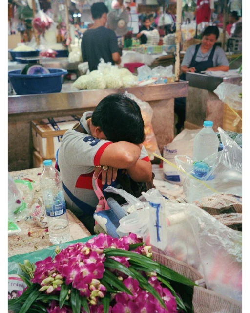 Man resting at a market stall surrounded by purple flowers, water bottles, and clear plastic bags, with other vendors in background.