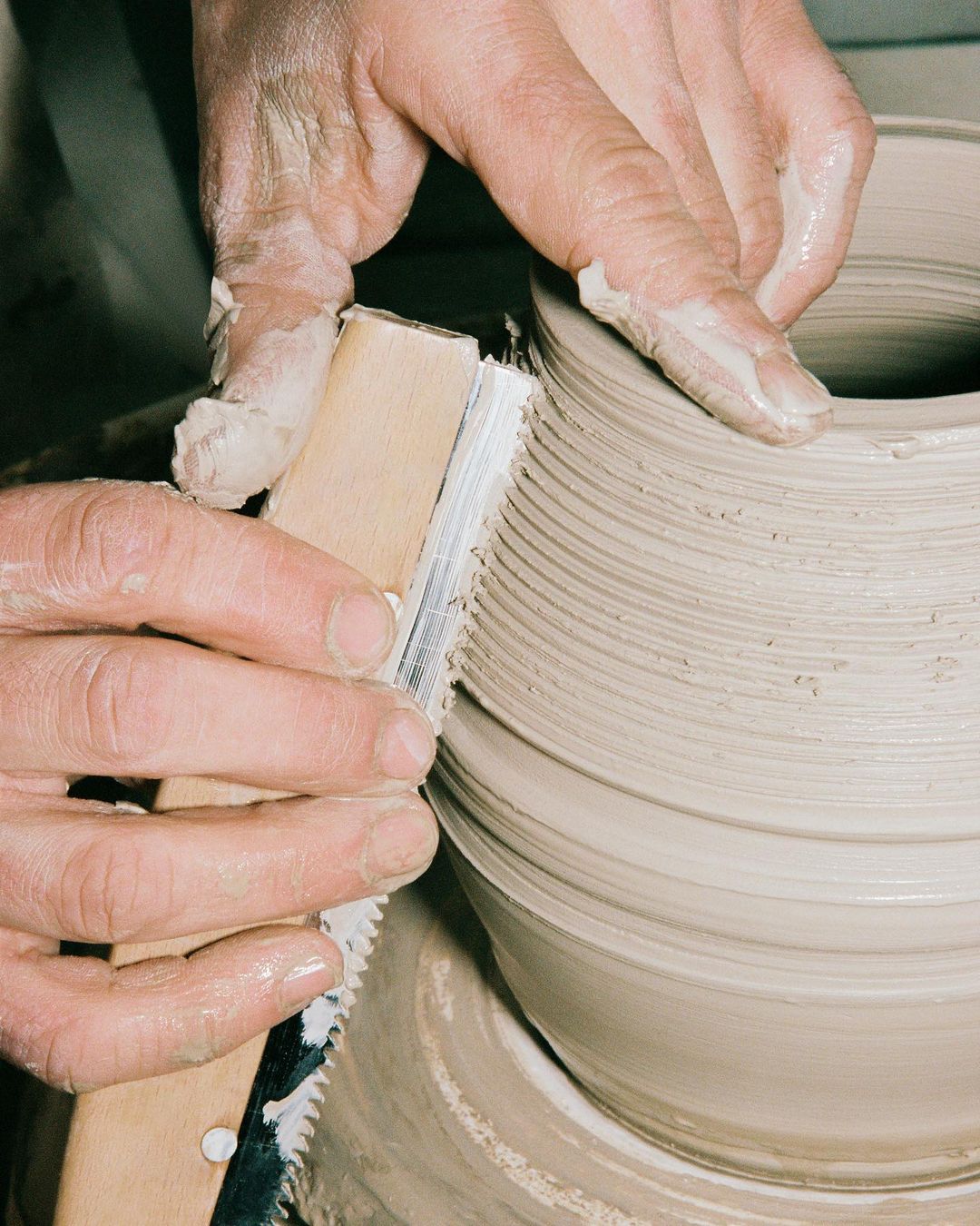 Human hands shaping clay on a pottery wheel using a shaping tool, creating a symmetrical form in a studio setting.