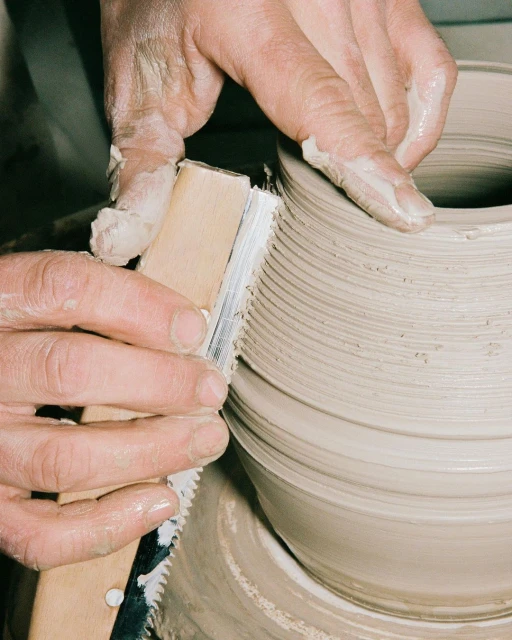 Human hands shaping clay on a pottery wheel using a shaping tool, creating a symmetrical form in a studio setting.