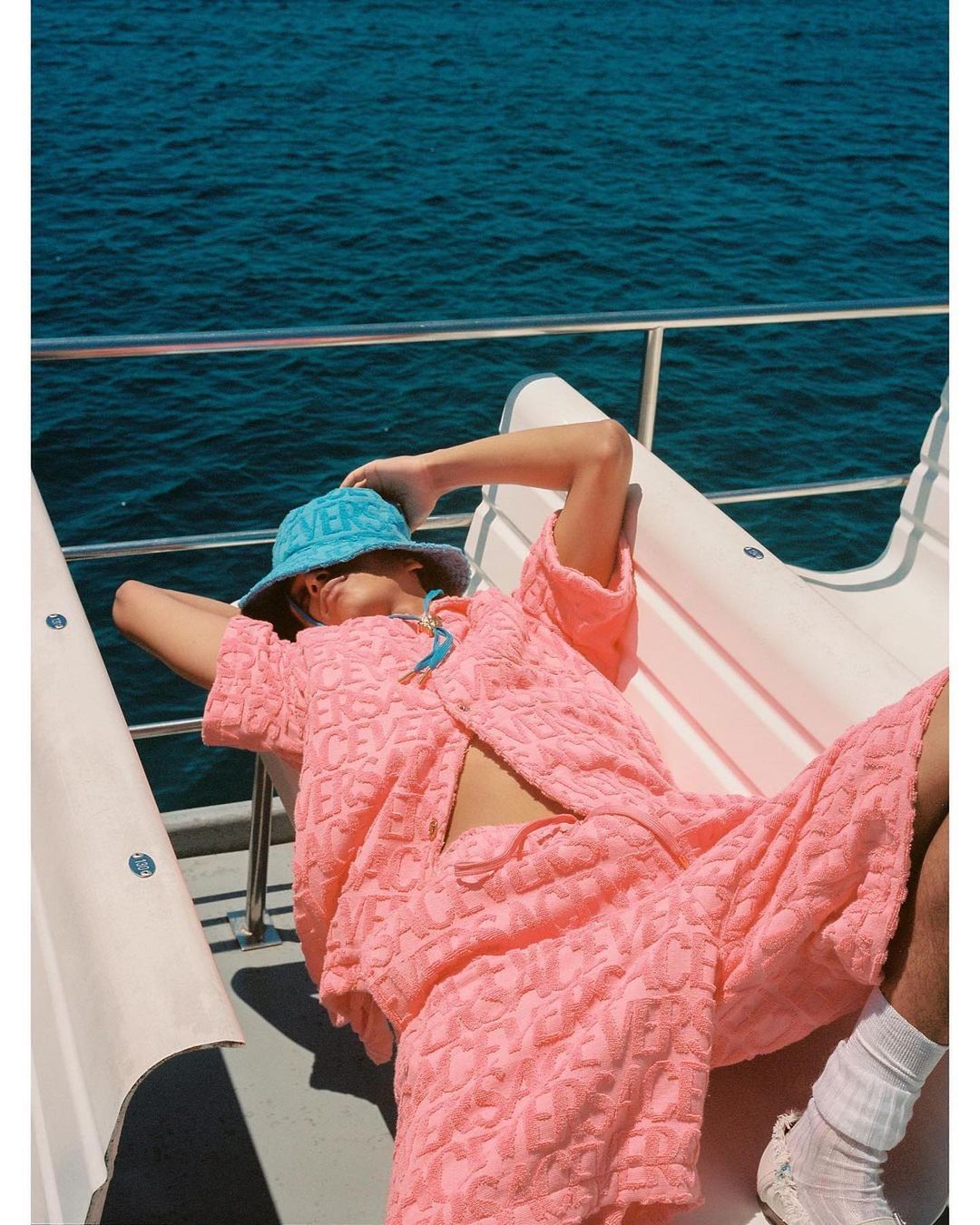 Person in pink textured co-ord set and light blue bucket hat relaxing on boat with blue ocean in background on sunny day.