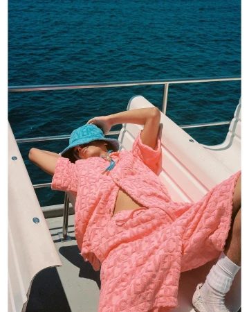 Person in pink textured co-ord set and light blue bucket hat relaxing on boat with blue ocean in background on sunny day.