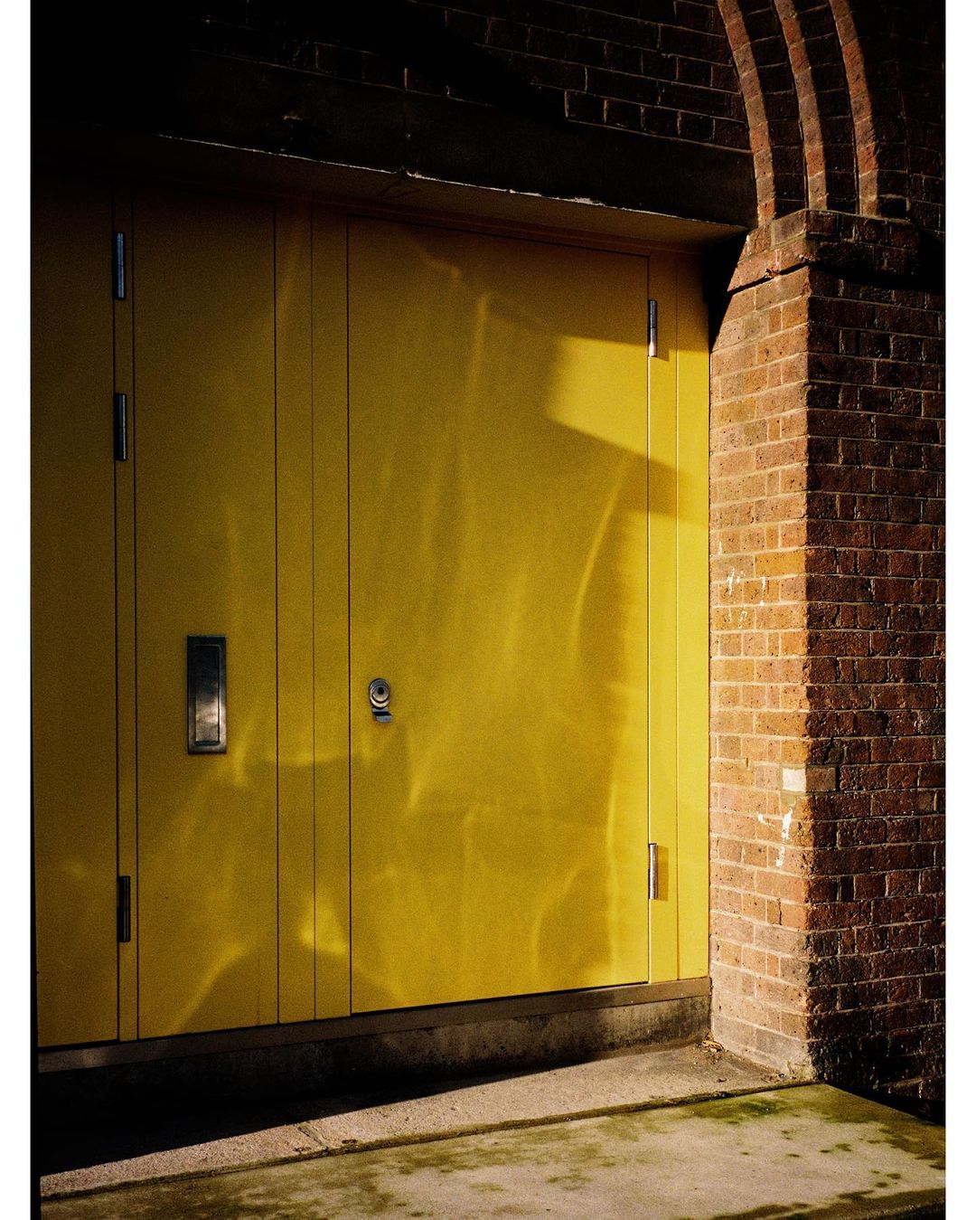 Exterior shot of a yellow door set in a red brick archway, featuring strong side lighting and shadows on the concrete base.