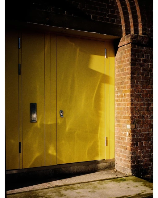 Exterior shot of a yellow door set in a red brick archway, featuring strong side lighting and shadows on the concrete base.