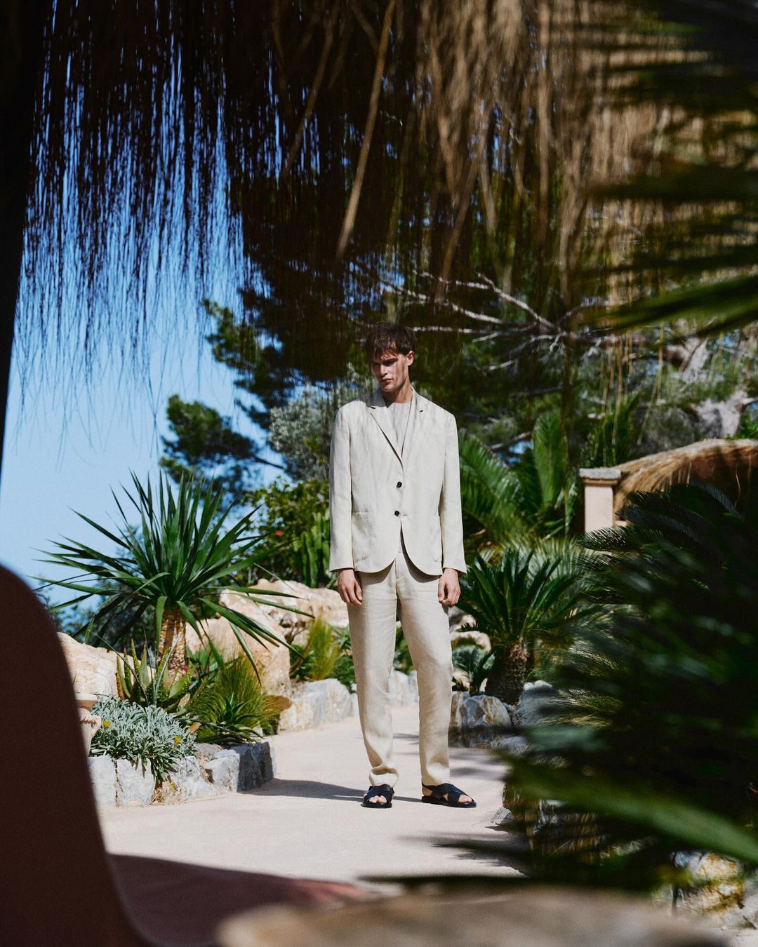 A male model in a beige suit poses in a sunlit outdoor scene with lush green plants and clear blue sky, showcasing high-end fashion.
