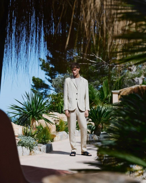 A male model in a beige suit poses in a sunlit outdoor scene with lush green plants and clear blue sky, showcasing high-end fashion.