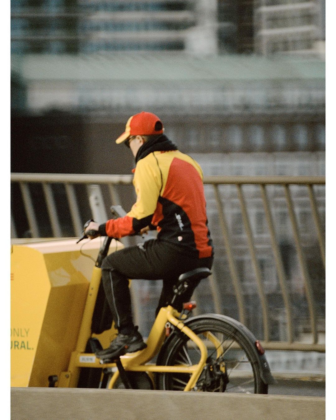 Delivery person rides a yellow cargo bike along a city street with tall buildings in the background, wearing a red and yellow jacket.