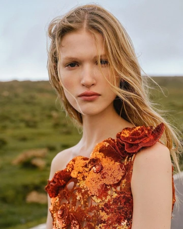 Blonde woman poses outdoors in an orange, floral-embellished dress against a blurred green and cloudy sky backdrop in a medium shot.