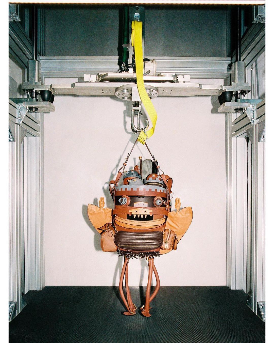 Steampunk-style robot-shaped handbag suspended from a metal frame by yellow straps in front of a neutral wall in a studio setup.