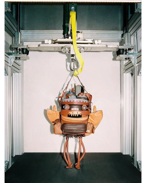 Steampunk-style robot-shaped handbag suspended from a metal frame by yellow straps in front of a neutral wall in a studio setup.