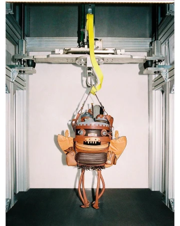 Steampunk-style robot-shaped handbag suspended from a metal frame by yellow straps in front of a neutral wall in a studio setup.
