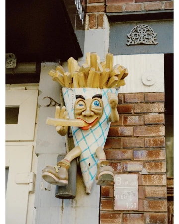 Sculpture of a cartoon-like figure made of french fries and a paper cone, mounted on the outside wall of a brick building.