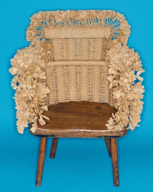 Rustic wooden chair with woven wicker back, decorated with paper flowers, presented against a bright blue background in a studio shot.