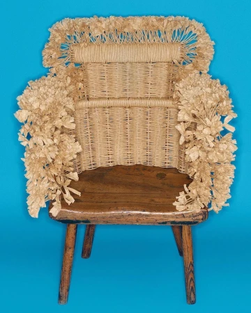 Rustic wooden chair with woven wicker back, decorated with paper flowers, presented against a bright blue background in a studio shot.