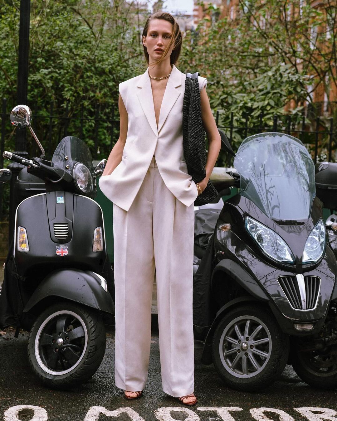 Woman in a sleeveless blazer and wide-leg trousers stands between two scooters, blending fashion with urban transport.