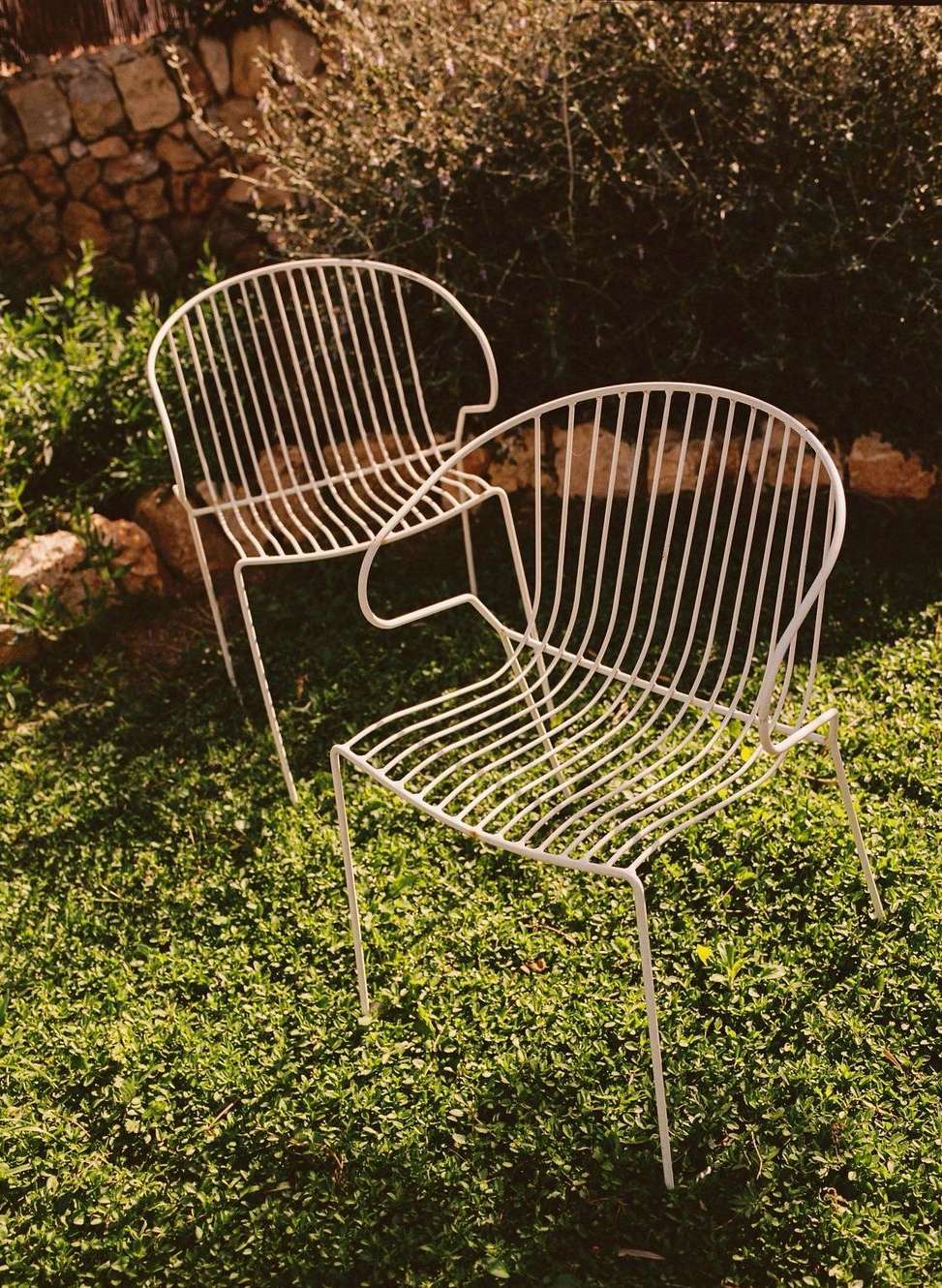 Two minimalist white wire chairs sit on a lush green lawn in an outdoor setting, bathed in natural light with a stone wall backdrop.