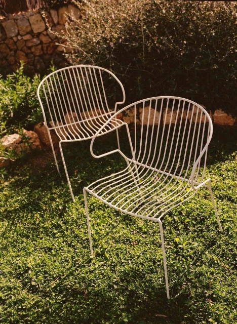 Two minimalist white wire chairs sit on a lush green lawn in an outdoor setting, bathed in natural light with a stone wall backdrop.