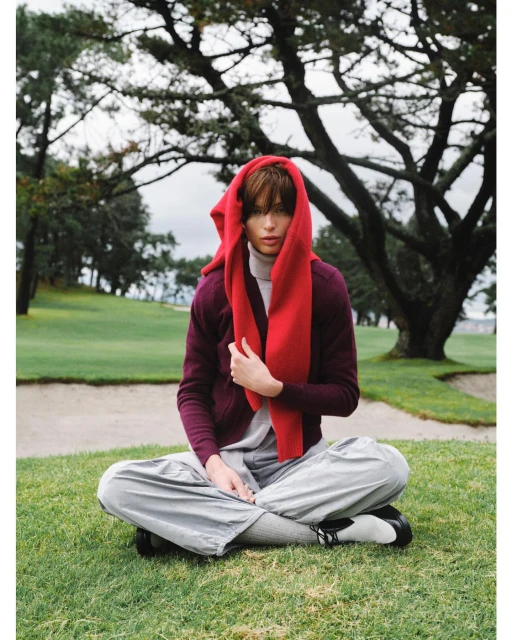 Female model sits cross-legged on green grass in gray pants and a red scarf-hood, with trees and a cloudy sky behind her.
