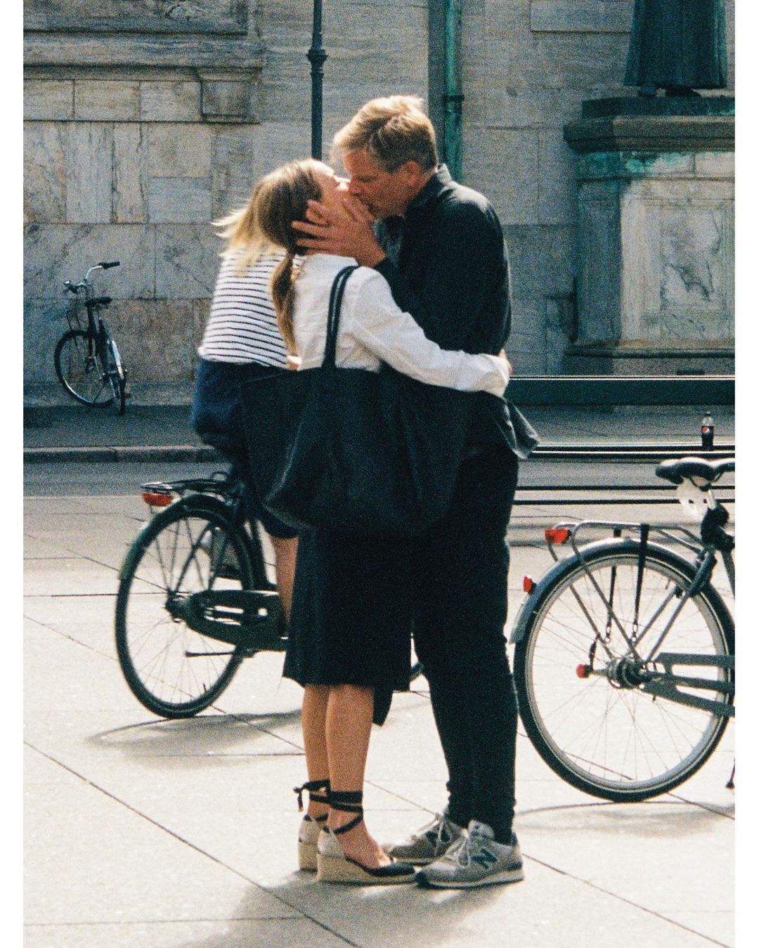 A man and woman kissing on a public square outdoors next to parked bicycles, photographed by Yago Castromil.