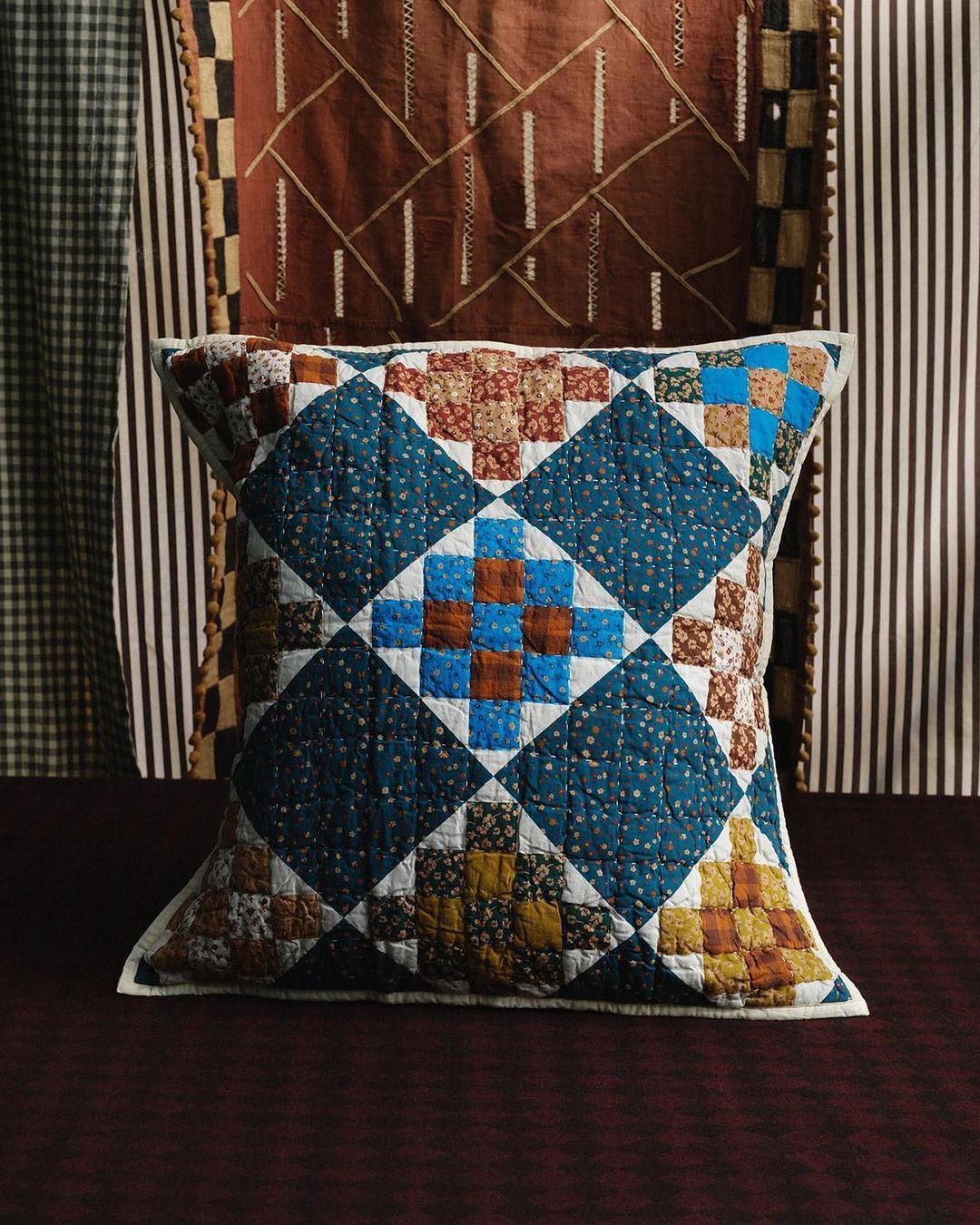 A cozy quilted pillow features blue and brown patterns in a home interior setting with striped and geometric-patterned textiles in the background.