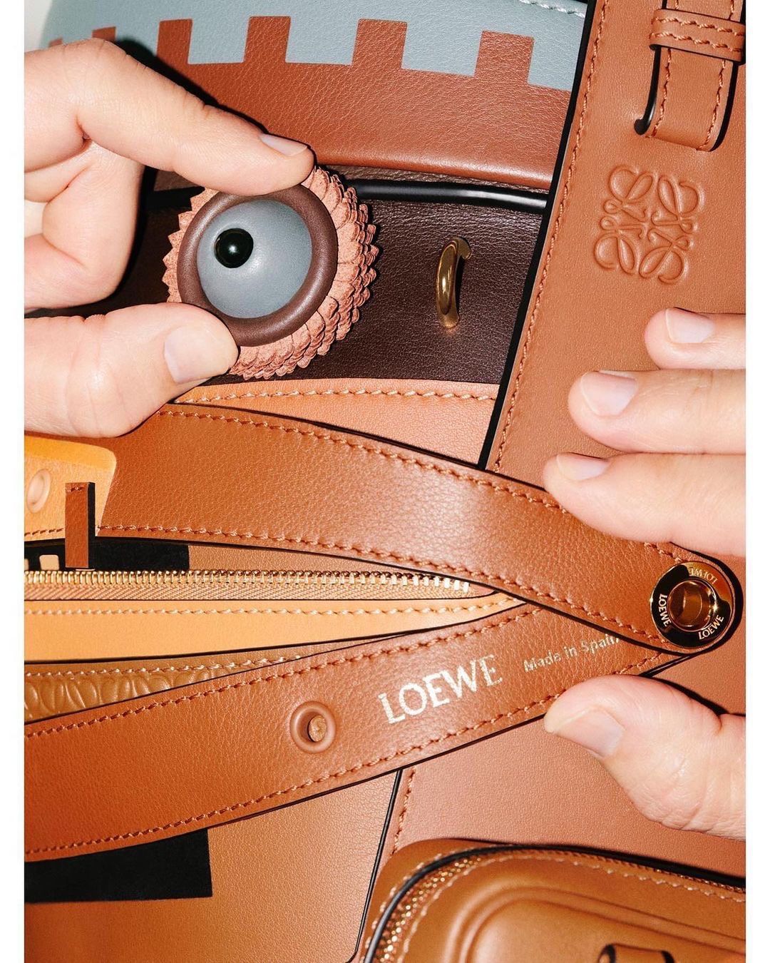 Close-up of a brown Loewe handbag with a cartoonish eye attached, held by a hand, showcasing the brand's logo and design details.