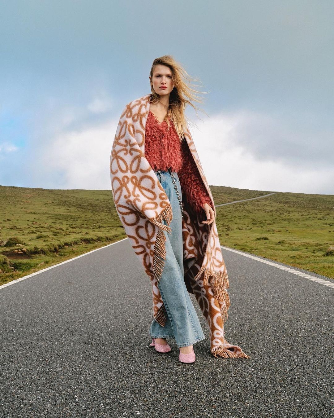Model standing on a paved road wearing a patterned blanket, red textured top, wide leg denim jeans, and pink shoes.