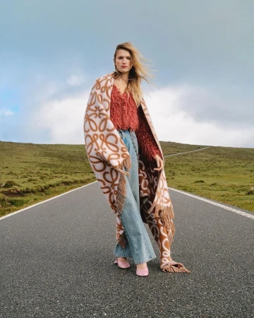 Model standing on a paved road wearing a patterned blanket, red textured top, wide leg denim jeans, and pink shoes.