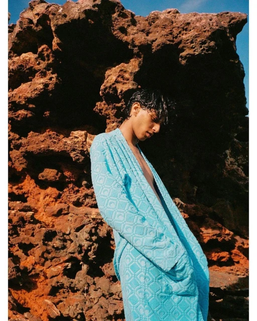 A person wearing a blue robe poses in front of a large, reddish-brown rock formation outdoors with a partly cloudy sky above.