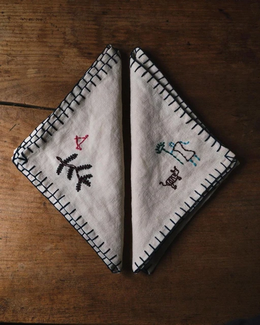 Two embroidered fabric napkins with black stitching and simplistic tree and animal patterns on a wooden table.