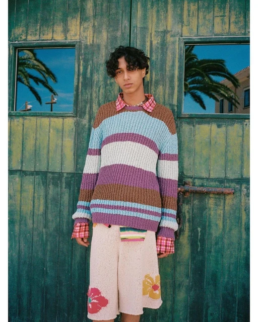 Young adult stands against weathered green doors wearing a striped knit sweater and floral-embroidered shorts, gazing at the camera.