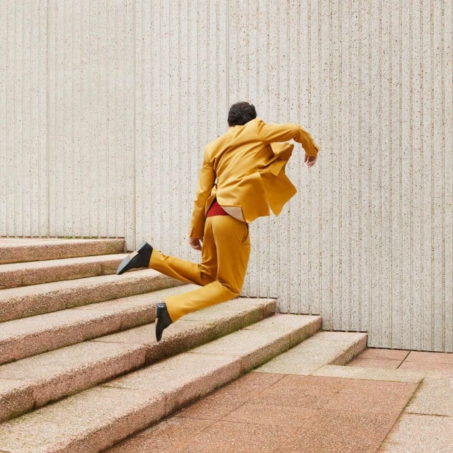 An adult in a mustard-colored suit jumps up a set of concrete stairs with a textured wall in the background on an overcast day.