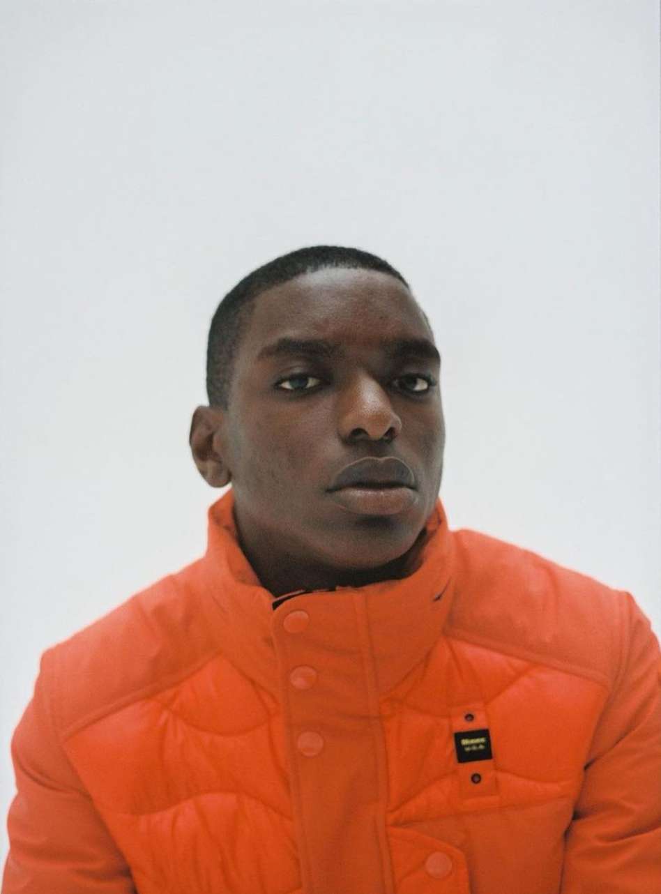 Portrait of a young adult male of African descent wearing an orange puffer jacket, set against a bright white background in a studio.