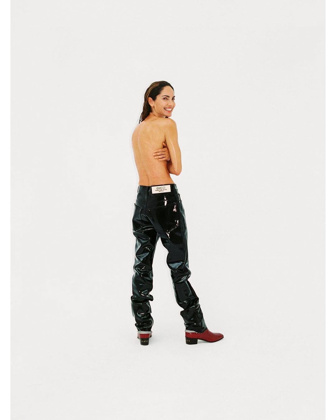 A slim adult woman smiles over her shoulder, posing topless in shiny black pants and reddish-brown boots against a white backdrop.