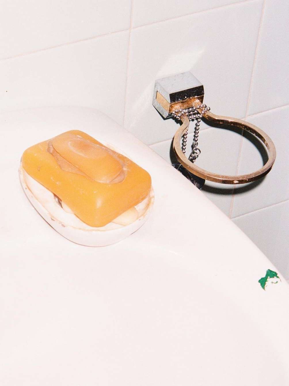 Close-up studio shot of a bar of orange soap in a white soap dish next to a metal soap holder on a tiled white wall.